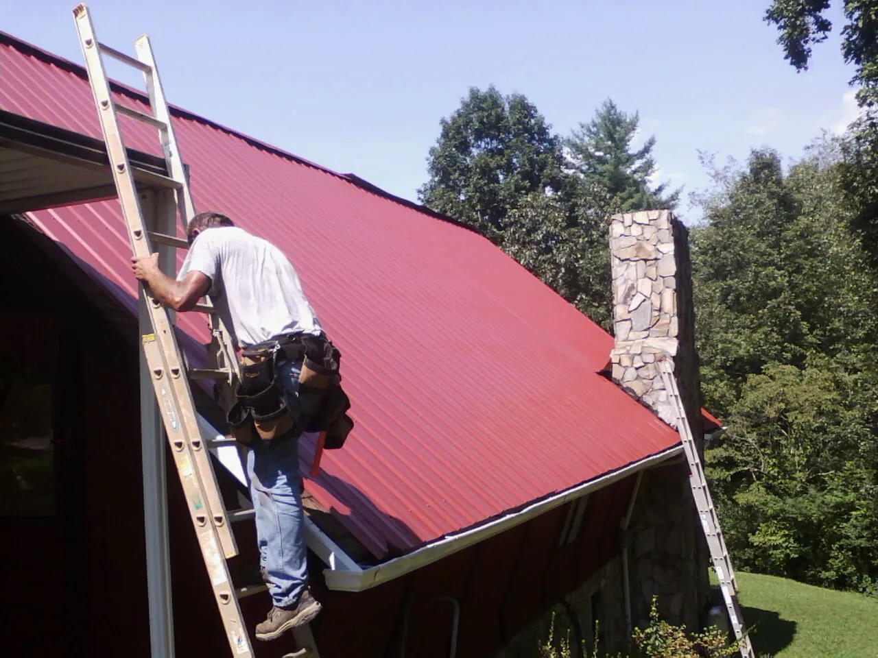 Roofer on a ladder beside a red metal roof and stone chimney — Soffit & Fascia Repair by Roofing Brothers in Forest Grove